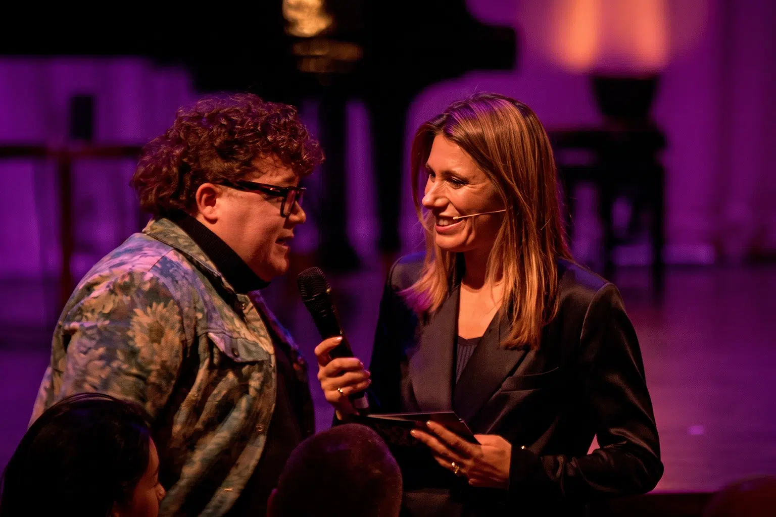 Twee personen in gesprek op een podium tijdens een live evenement met paarse verlichting.