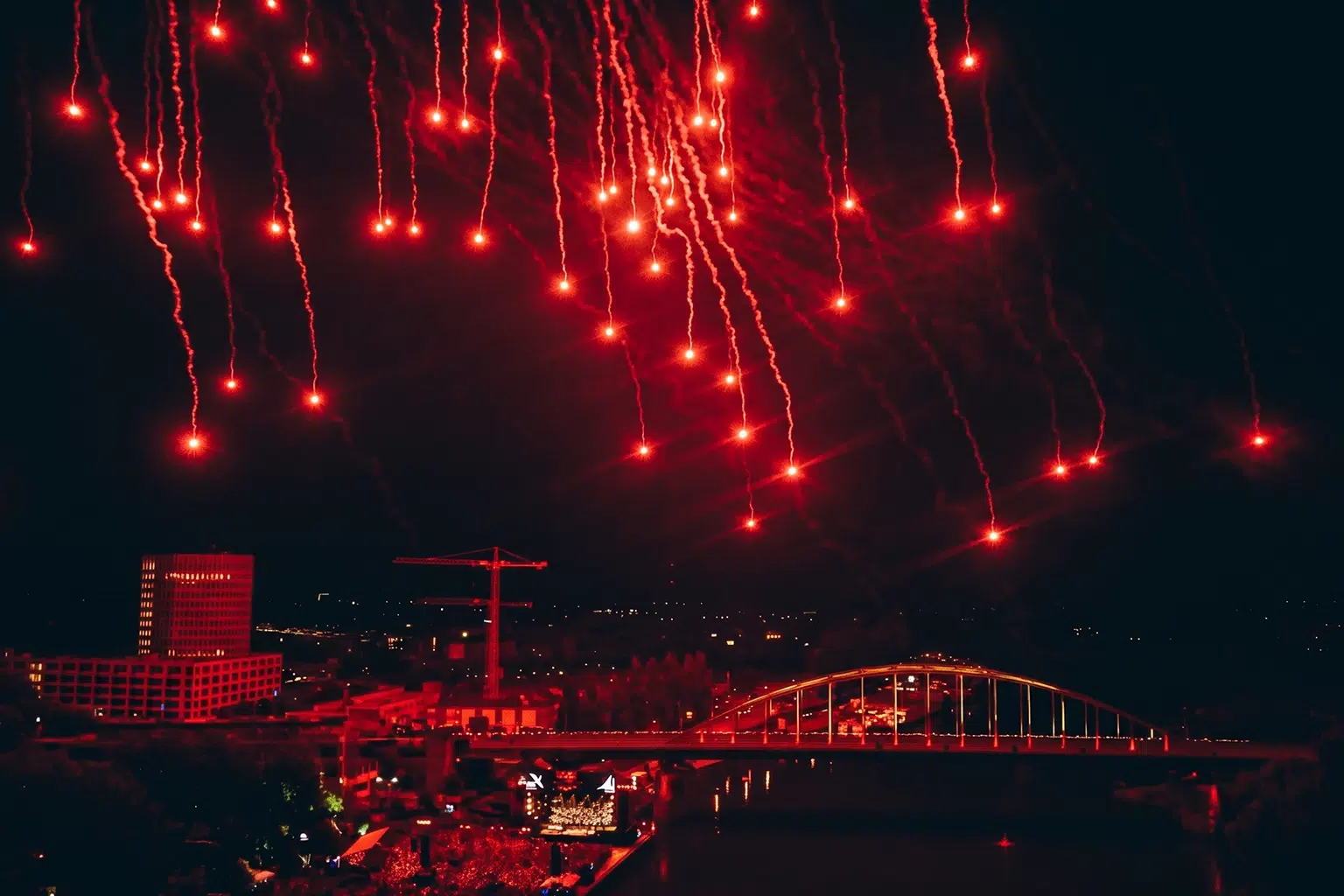 Rood vuurwerk boven een stad met brug en publiek langs het water.